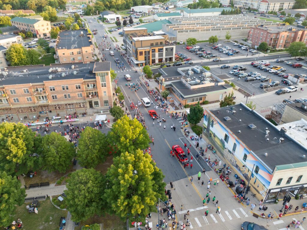 Aerial view of a small town street parade with people gathered on sidewalks, several vehicles in the procession, and buildings and parking lots surrounding the scene.