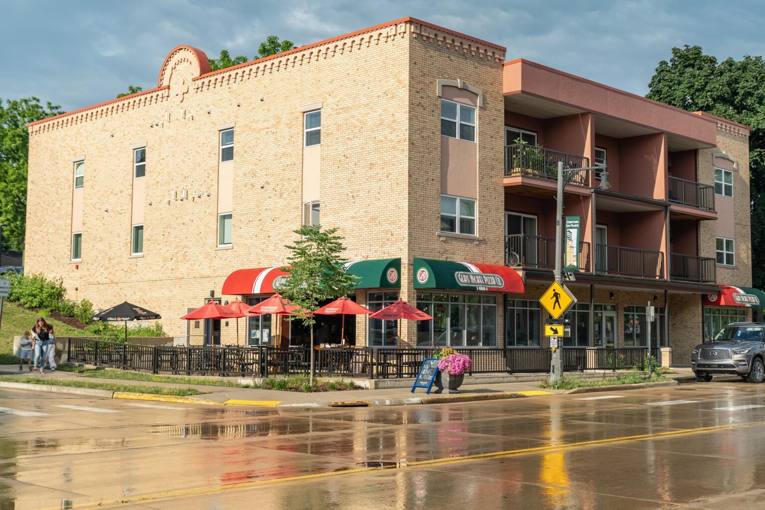 A brick building with ground-floor restaurant, red umbrellas on the outdoor patio, upper-level balconies, and wet streets reflecting the scene.