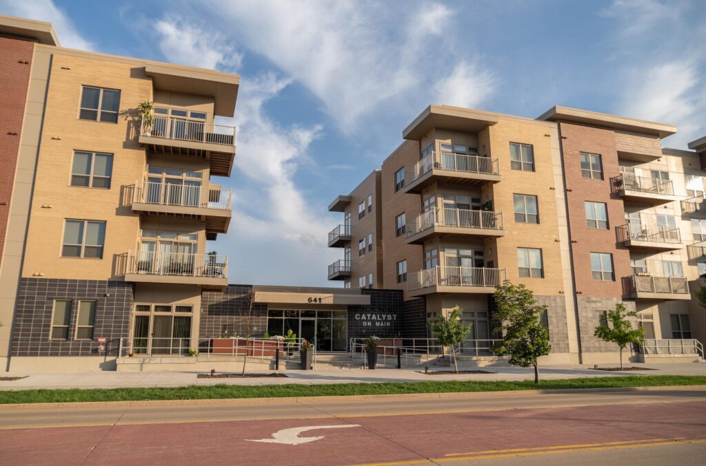 Modern four-story apartment buildings with balconies and large windows, separated by an entrance labeled "Catalyst on Main." Trees and a sidewalk line the street in front.