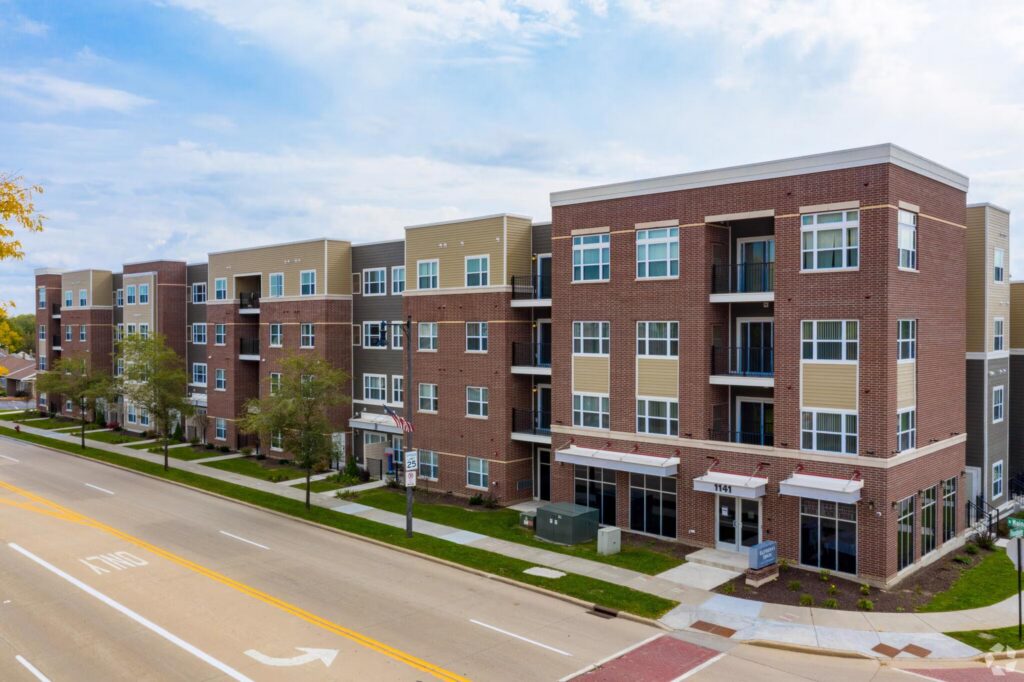 A modern three-story apartment building with brick and tan siding is situated along a wide street with trees, sidewalks, and parked cars.