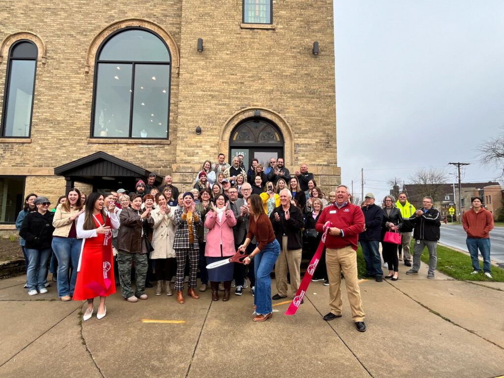 A group of people gathered outside a brick building, smiling and celebrating as a woman cuts a large ribbon during a ribbon-cutting ceremony.