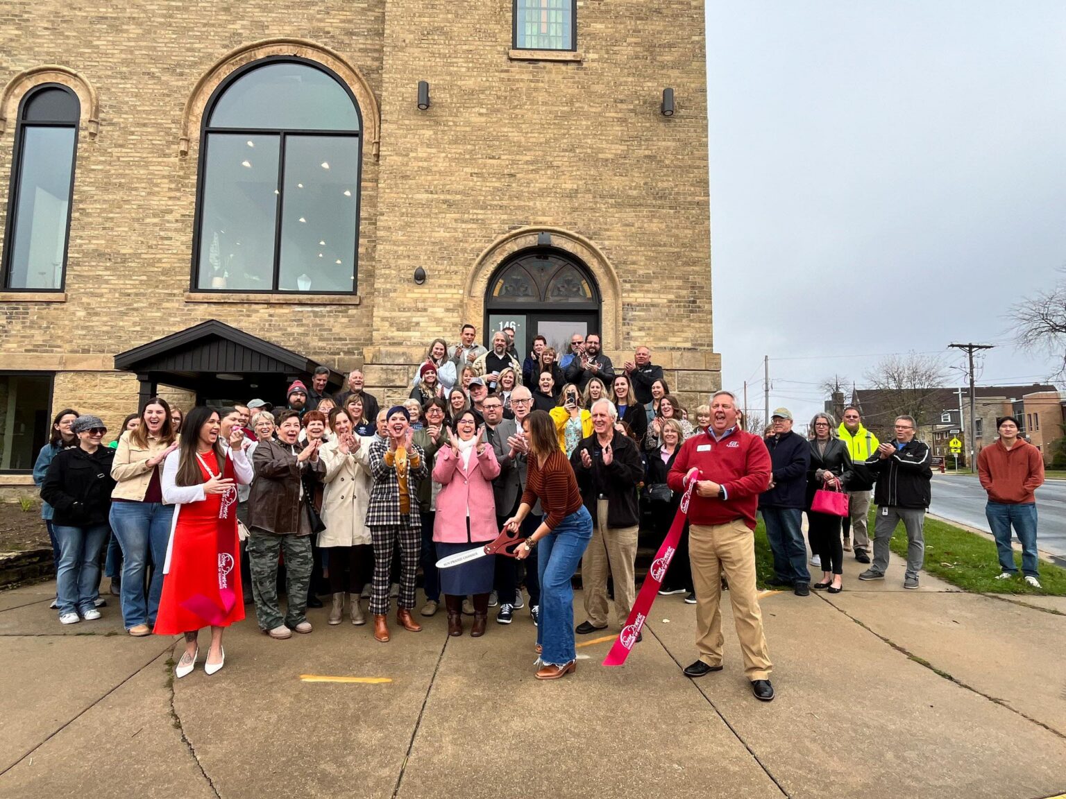 A group of people gathered outside a brick building, smiling and celebrating as a woman cuts a large ribbon during a ribbon-cutting ceremony.