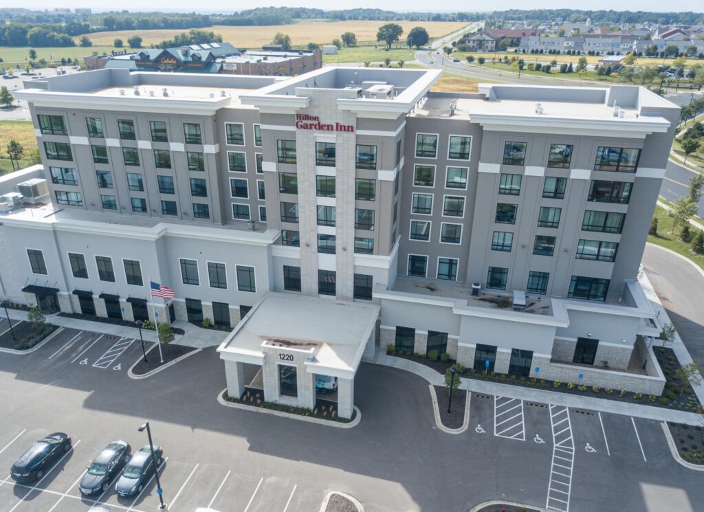 Aerial view of a Hilton Garden Inn hotel with multiple floors, a front entrance with a covered driveway, and a parking lot with marked spaces.