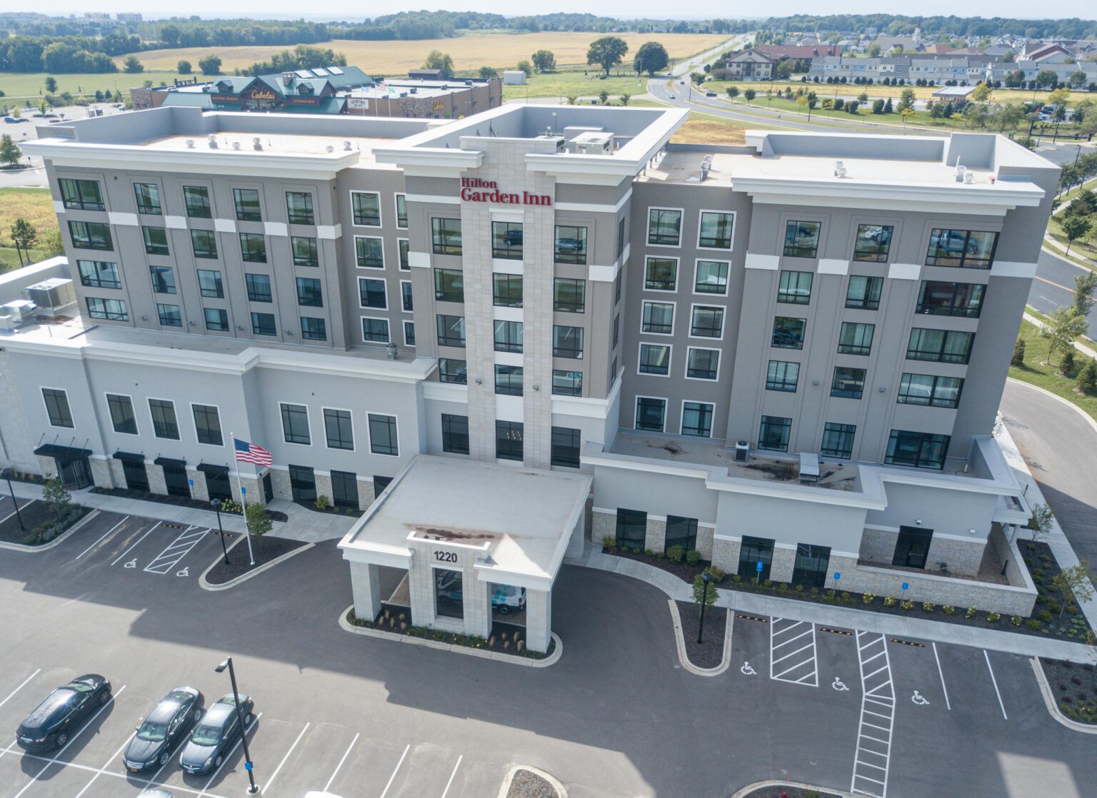 Aerial view of a Hilton Garden Inn hotel with multiple floors, a front entrance with a covered driveway, and a parking lot with marked spaces.