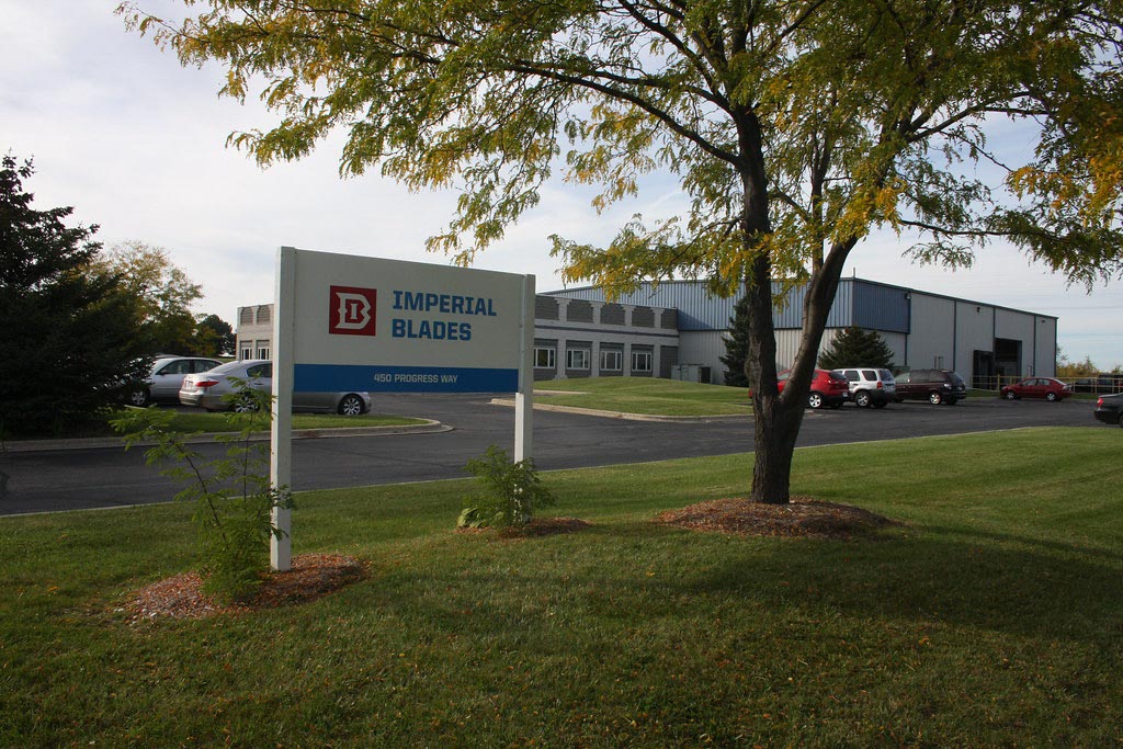 A sign for Imperial Blades stands on a grassy lawn in front of a commercial building with several cars parked outside.