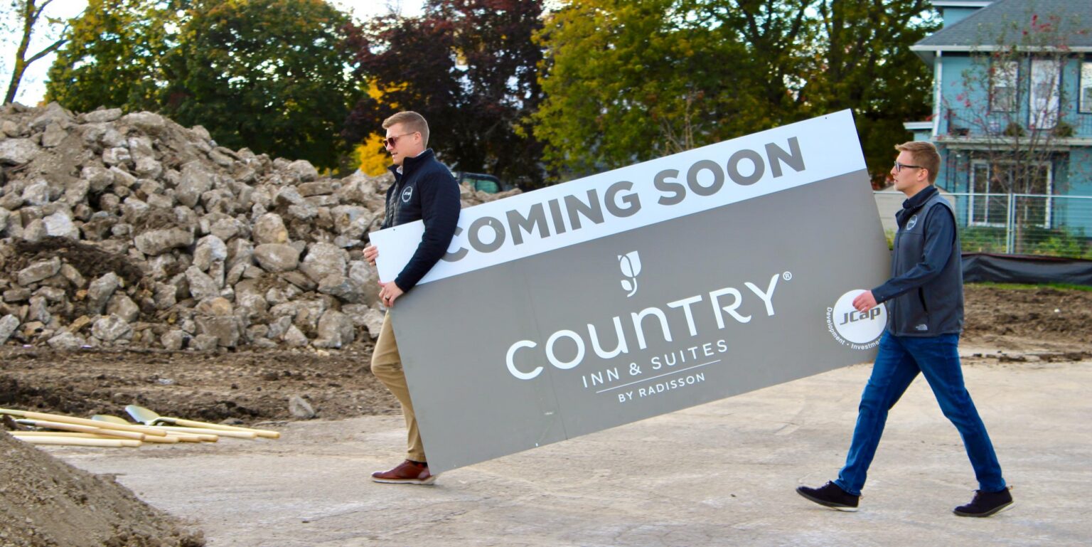 Two people carry a large “Coming Soon Country Inn & Suites by Radisson” sign at a construction site with dirt, rocks, and trees in the background.