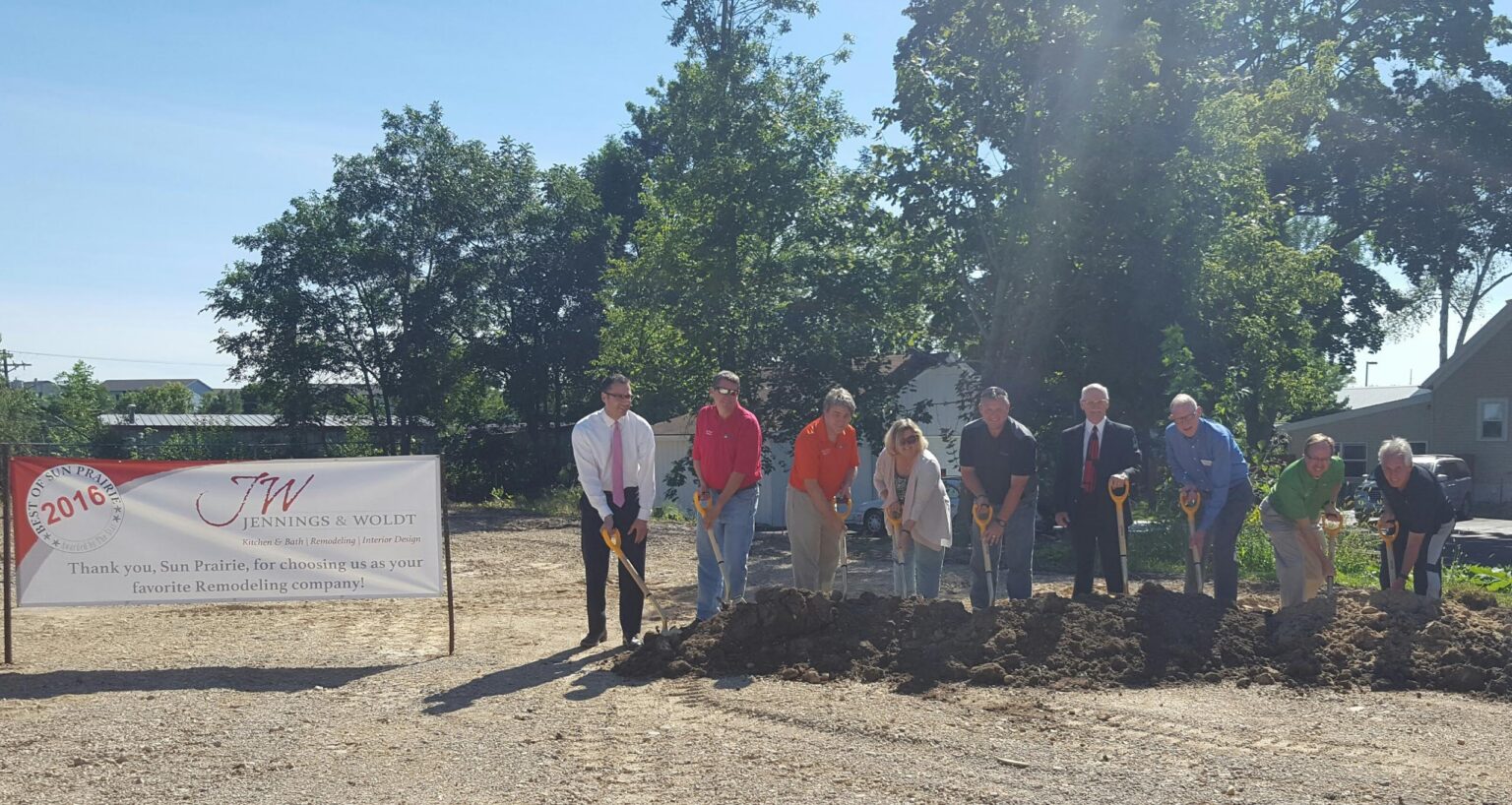 A group of people with shovels stand in a row at a groundbreaking ceremony next to a banner for Jennings & Woldt Remodeling, with trees and buildings in the background.