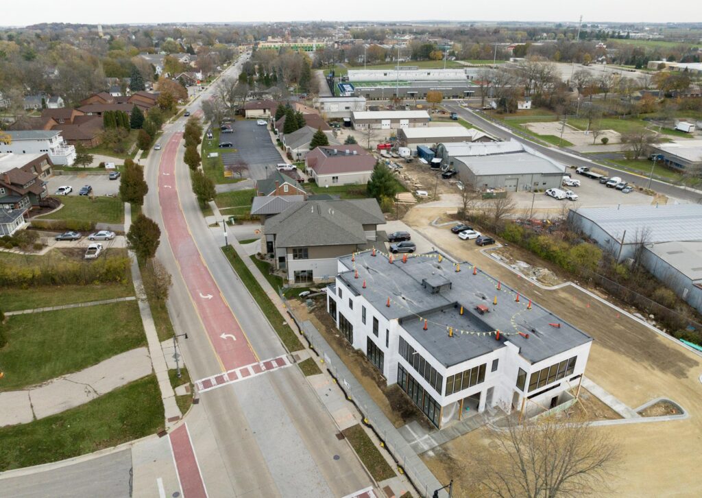 Aerial view of a suburban street with a newly constructed white building, nearby parking lots, and residential houses; overcast weather and sparse tree coverage.