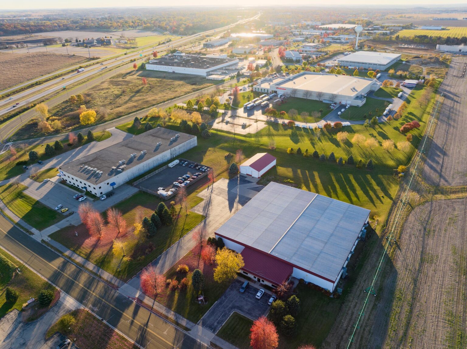Aerial view of an industrial park with multiple warehouses, parking lots, surrounding trees, and nearby roads during autumn.