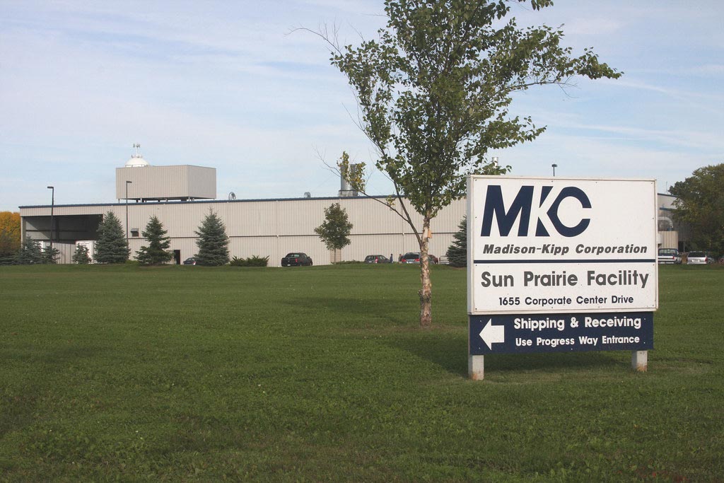 A large industrial building with a sign in front reading "MKC Madison-Kipp Corporation Sun Prairie Facility" and directions for shipping and receiving.