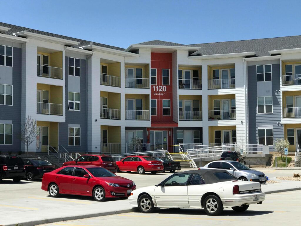 A modern three-story apartment building with balconies, labeled "1120 Building 1," and several parked cars in the foreground.