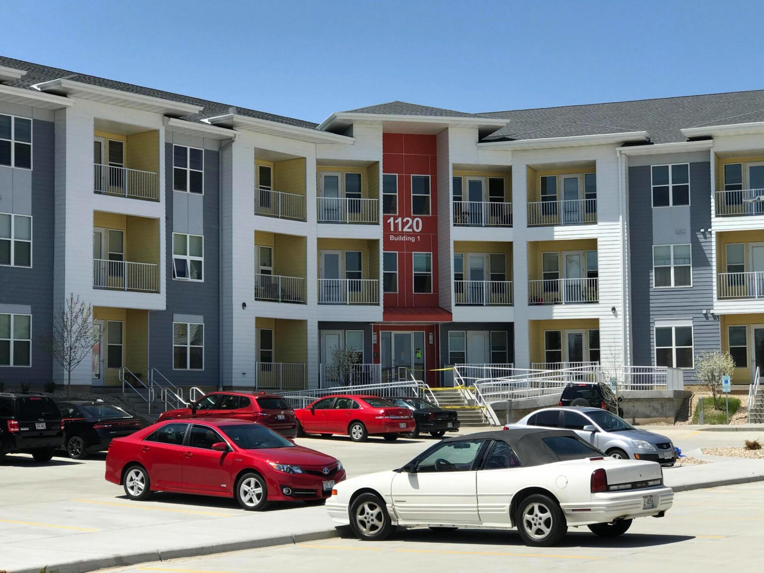 A modern three-story apartment building with balconies, labeled "1120 Building 1," and several parked cars in the foreground.