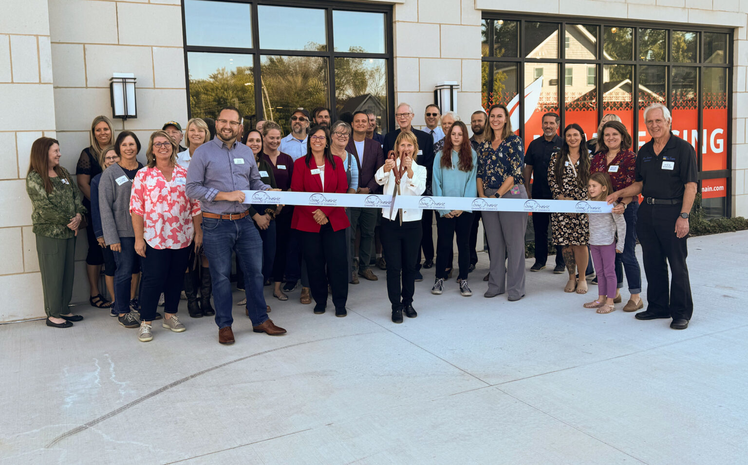 A group of people stands outside a building as a woman in the center cuts a ceremonial ribbon, marking an official opening event.
