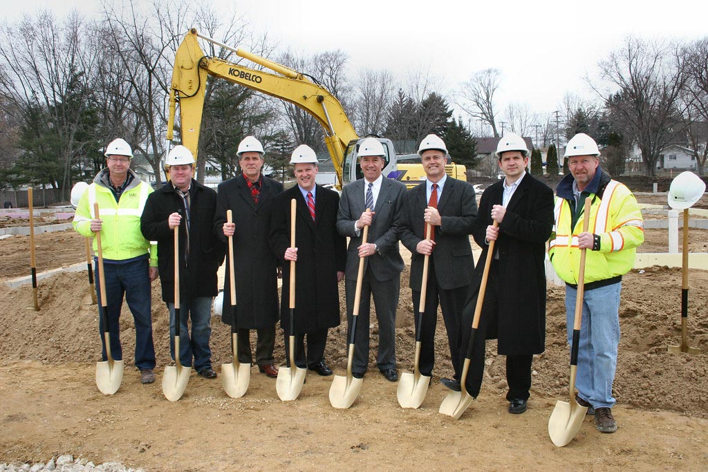 Eight men in hard hats and coats stand holding shovels at a construction site, with an excavator and building materials visible in the background.