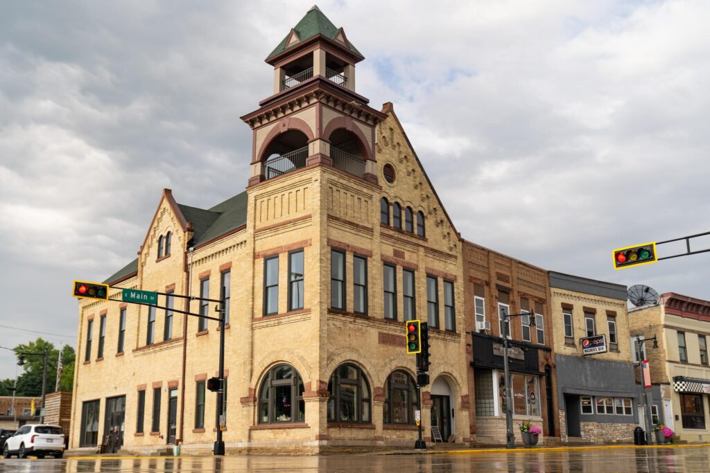 Historic brick building with arched windows and a prominent tower stands at a wet street corner under a cloudy sky, with traffic lights and small shops visible.