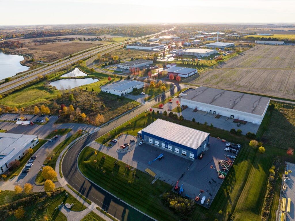 Aerial view of an industrial park with warehouses, parking lots, and roads surrounded by fields, trees, and a pond, taken during daylight.