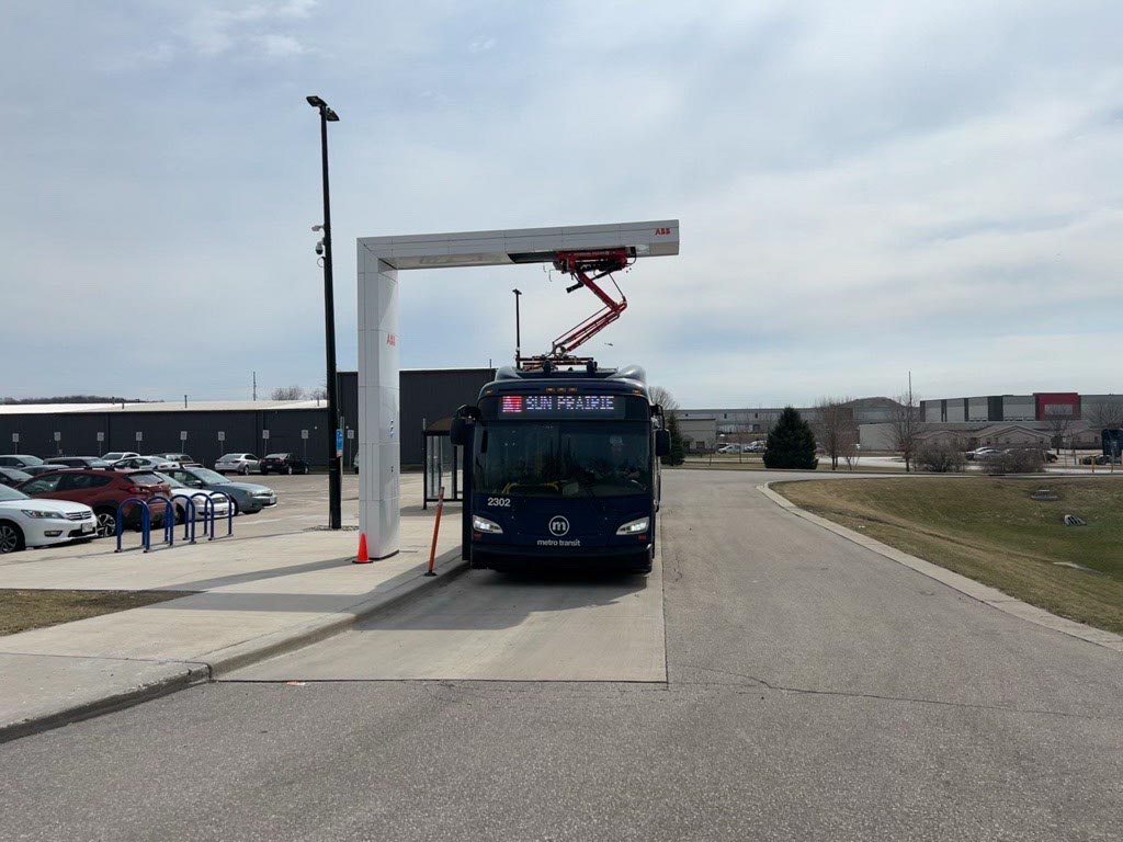 Electric bus stopped under a charging station arm in a parking lot, with cars parked to the side and industrial buildings in the background.