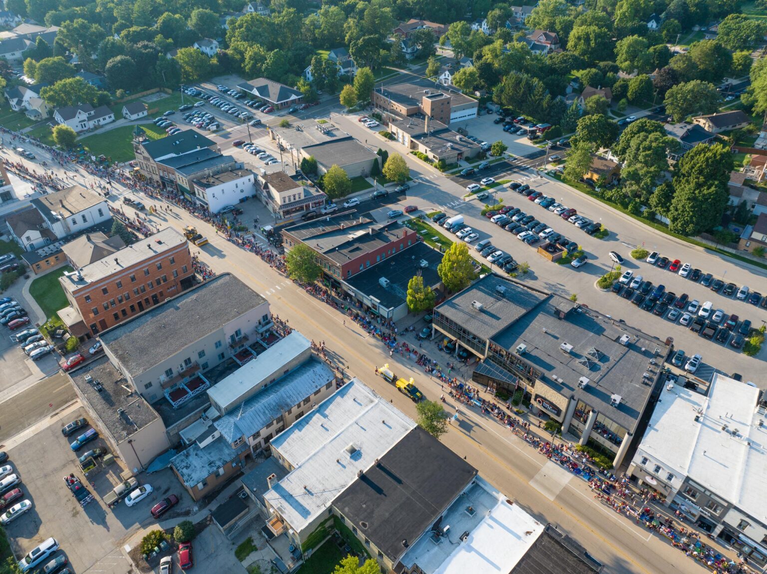 Aerial view of a town’s main street with a parade taking place; people line the sidewalks and cars are parked in lots and along the streets.