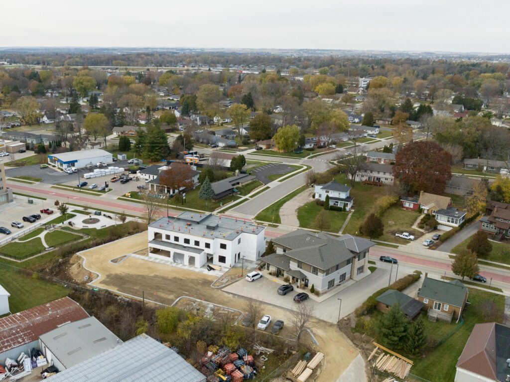 Aerial view of a suburban neighborhood with a construction site in the foreground, commercial buildings, houses, and tree-lined streets visible.