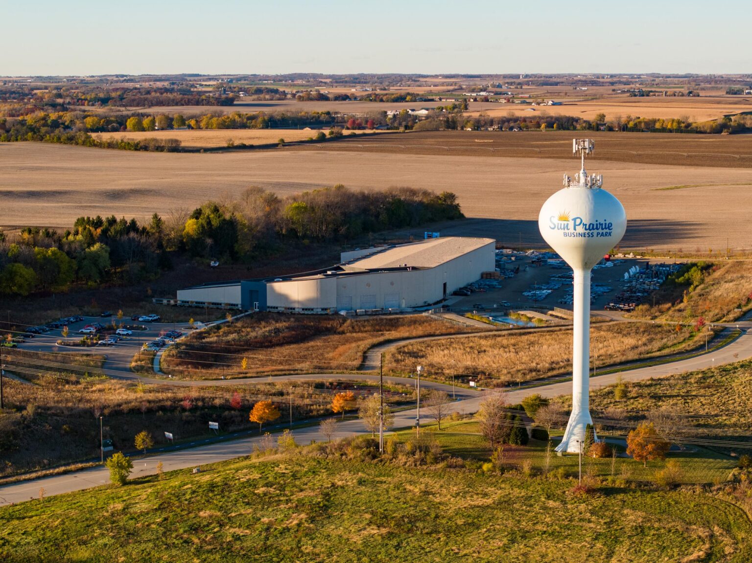 Aerial view of a water tower labeled "Sun Prairie Business Park" beside an industrial building and parking lot, surrounded by fields and trees in a rural landscape.