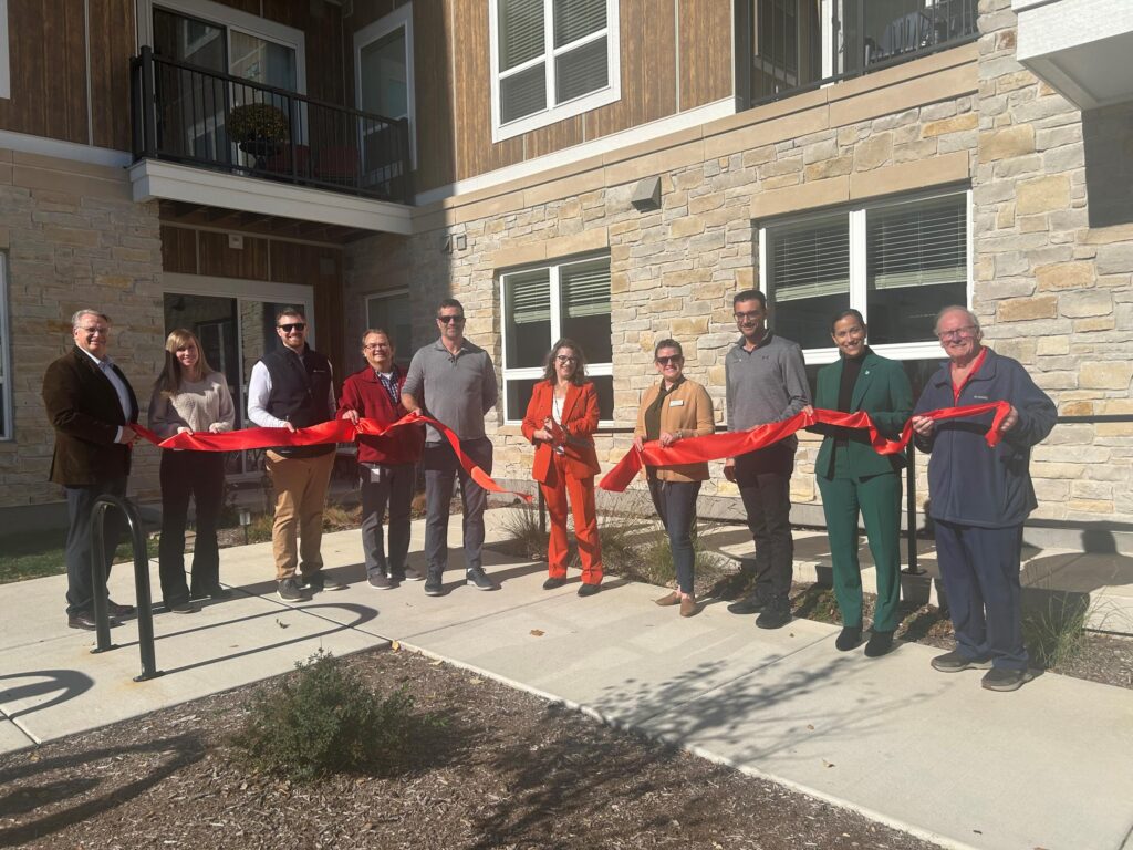 A group of people stand outside a building holding a red ribbon, preparing for a ribbon-cutting ceremony under sunny weather.