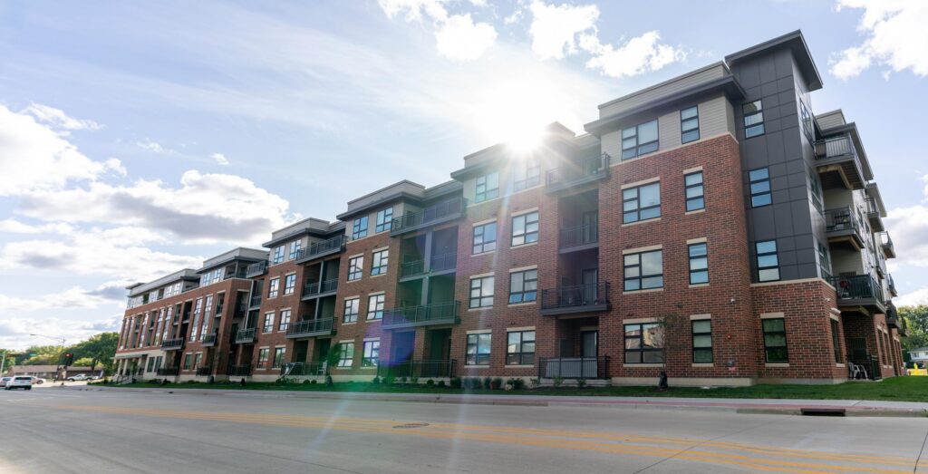A modern, multi-story brick apartment building with numerous windows and balconies, located along a wide street on a sunny day.