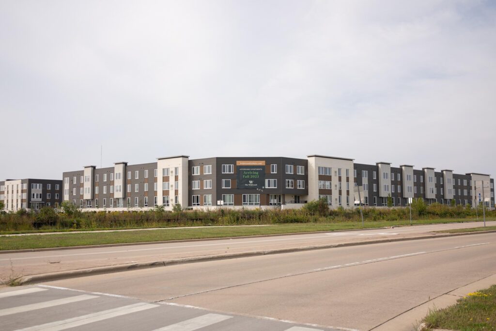 A modern three-story apartment complex with a sign reading "Now Leasing" is situated behind a grassy area and a wide paved road.