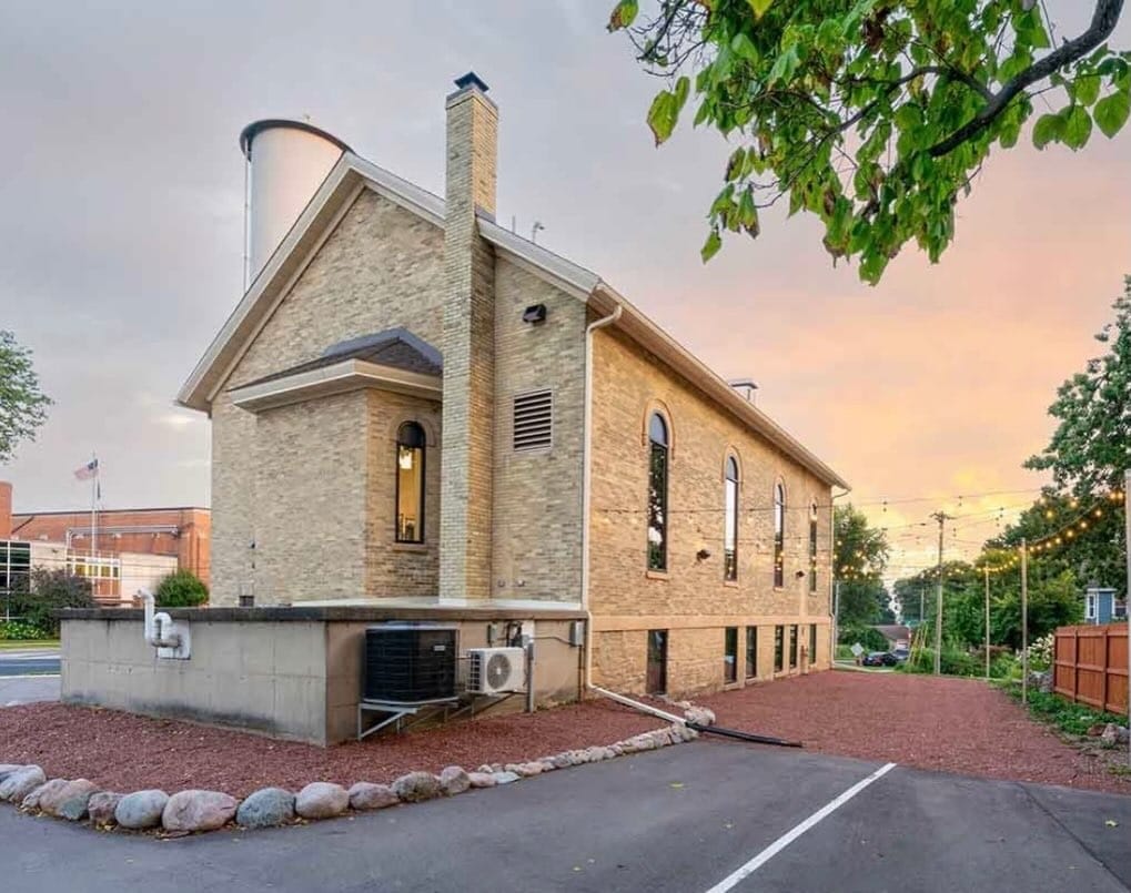 Two-story tan brick building with arched windows, a chimney, and HVAC units outside, photographed at sunset with a paved driveway and surrounding trees.