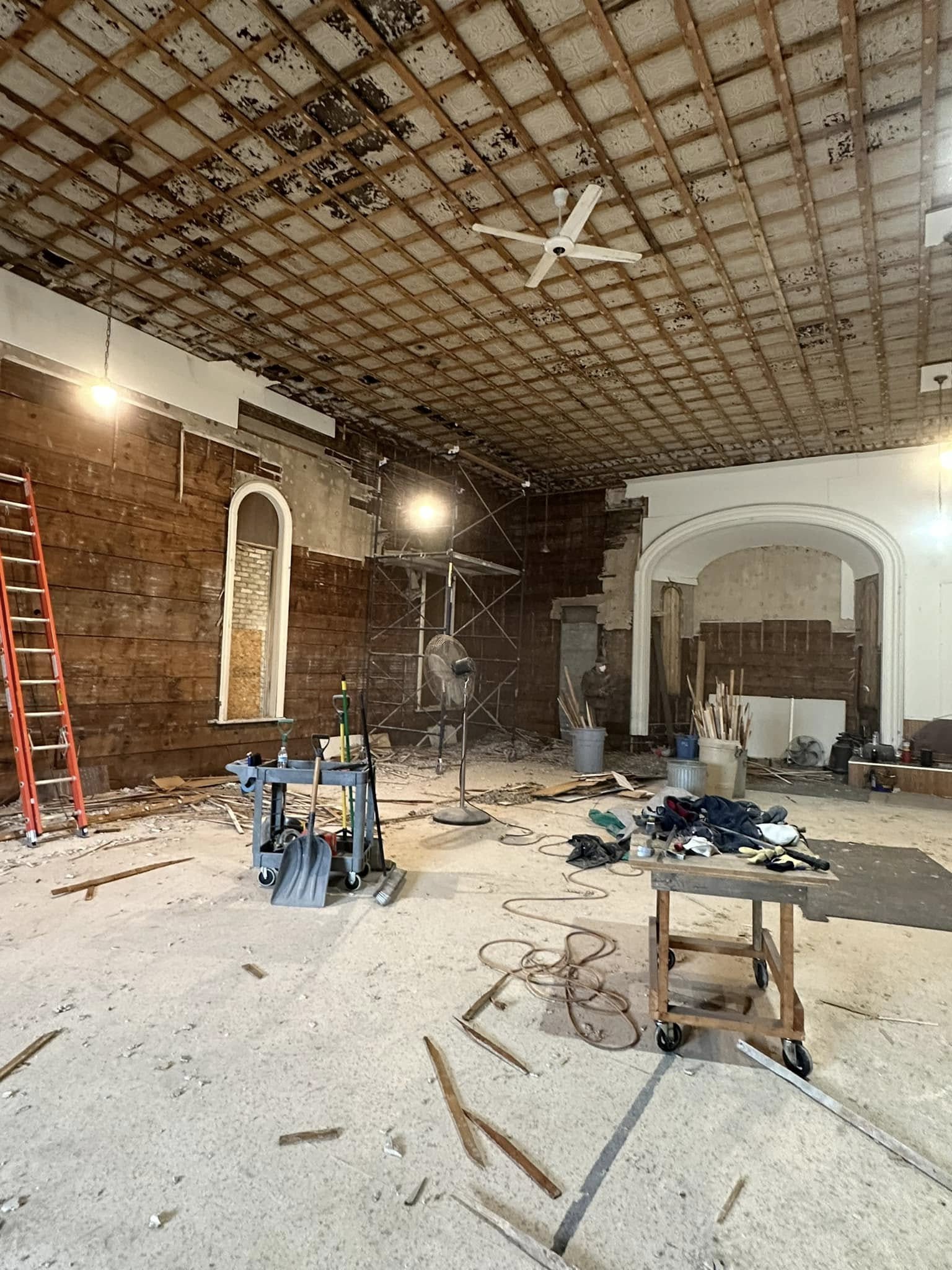 Large room under renovation with exposed ceiling grid, construction tools, scaffolding, a red ladder, and debris scattered across the floor. Two hanging lights provide illumination.