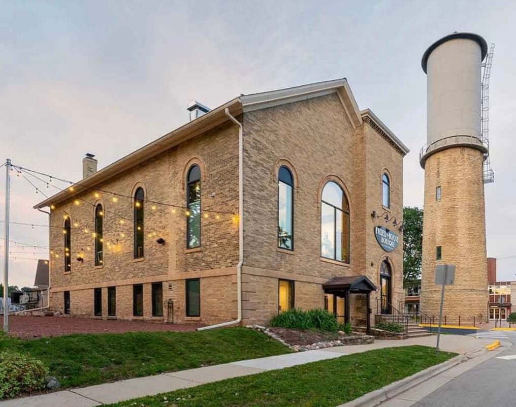 A two-story brick building with large arched windows next to a tall cylindrical water tower, with string lights outside and a sidewalk in front.