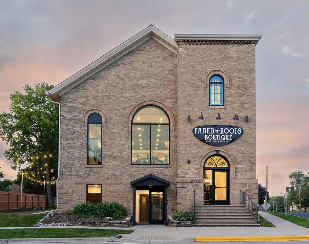 Two-story brick building with large arched windows and a sign reading “Faded Roots Boutique” on the front, surrounded by greenery and outdoor string lights.