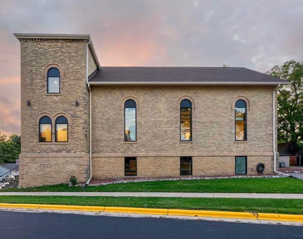 A tan brick building with five tall arched windows, minimal landscaping, and a sloped roof, seen from the side at sunset.