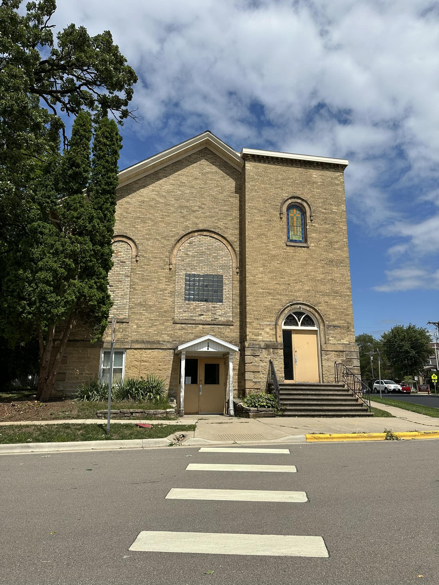 A two-story brick building with arched windows and two entrances, one with stairs, sits by a street with a crosswalk on a partly cloudy day.