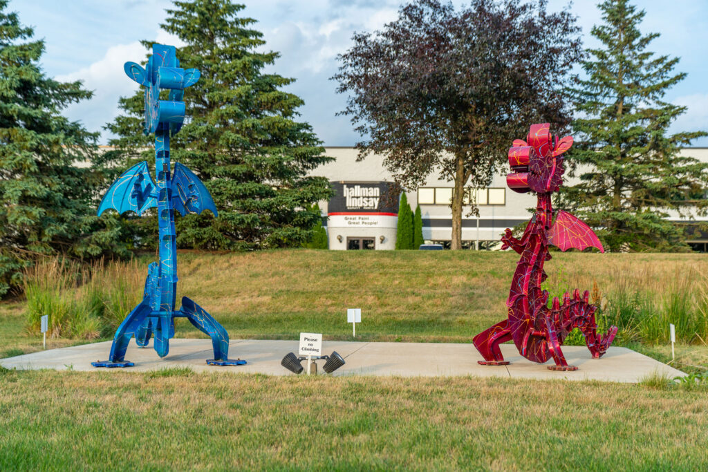 A blue metal dragon sculpture stands across from a red metal dragon sculpture on a concrete pad, with a “Please No Climbing” sign between them.