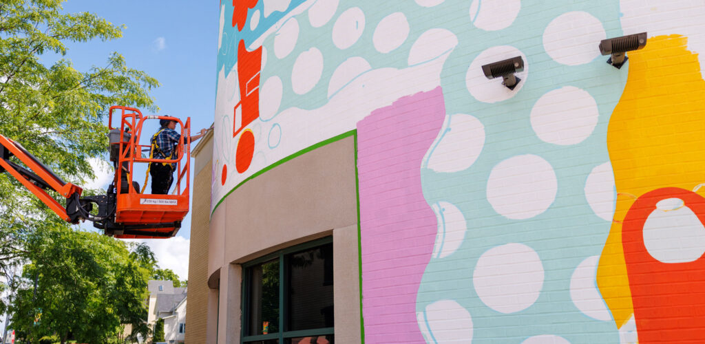 A person in a cherry picker paints a colorful geometric mural on the side of a brick building.