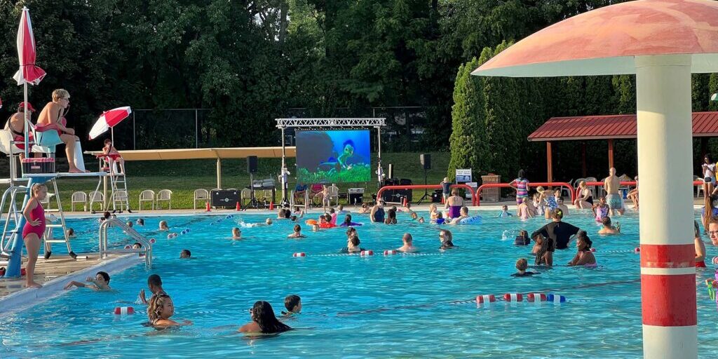 People swim and relax in a public outdoor pool while a large screen near the pool shows a movie; a lifeguard watches from a chair. Trees and shelters are in the background.