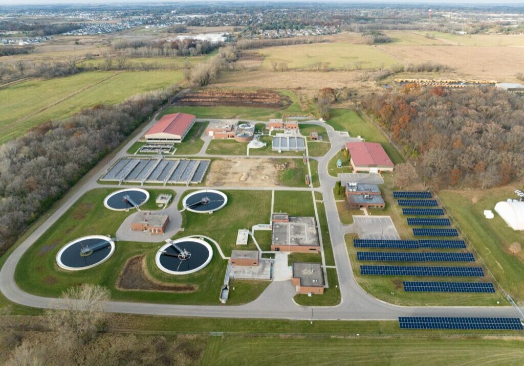 Aerial view of a wastewater treatment plant with several circular tanks, solar panel arrays, buildings, and access roads, surrounded by fields and trees.