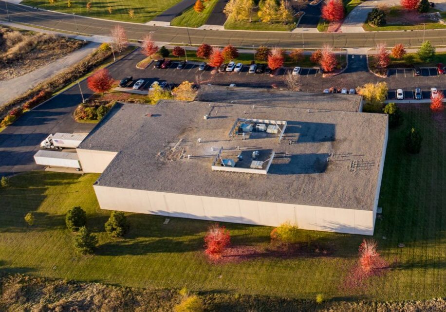 Aerial view of a large rectangular commercial building with a flat roof, surrounded by grass, parking lot, and trees with autumn foliage.