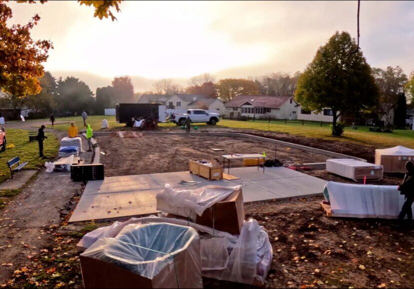 Construction workers and equipment on a residential building site with concrete foundation, materials, and houses in the background on a sunny morning.
