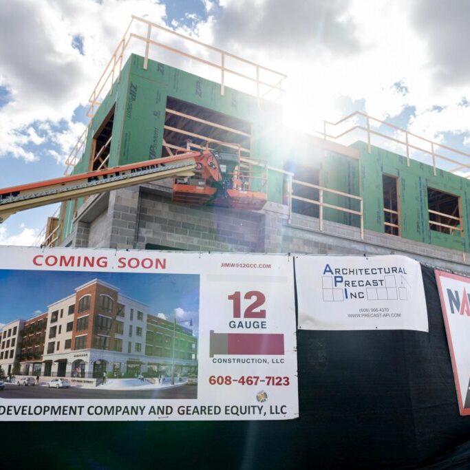 A multi-story building under construction with scaffolding and signs advertising leasing and upcoming availability. A lift and construction materials are visible.