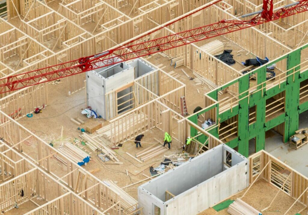 Aerial view of a building construction site with exposed wooden framing, scattered materials, workers, and a red crane overhead.