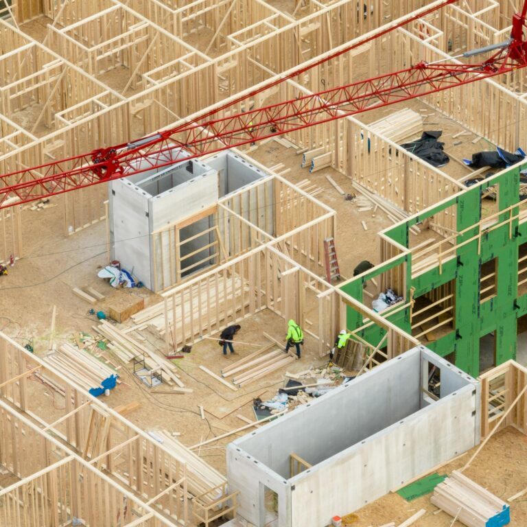 Aerial view of a building construction site with exposed wooden framing, scattered materials, workers, and a red crane overhead.