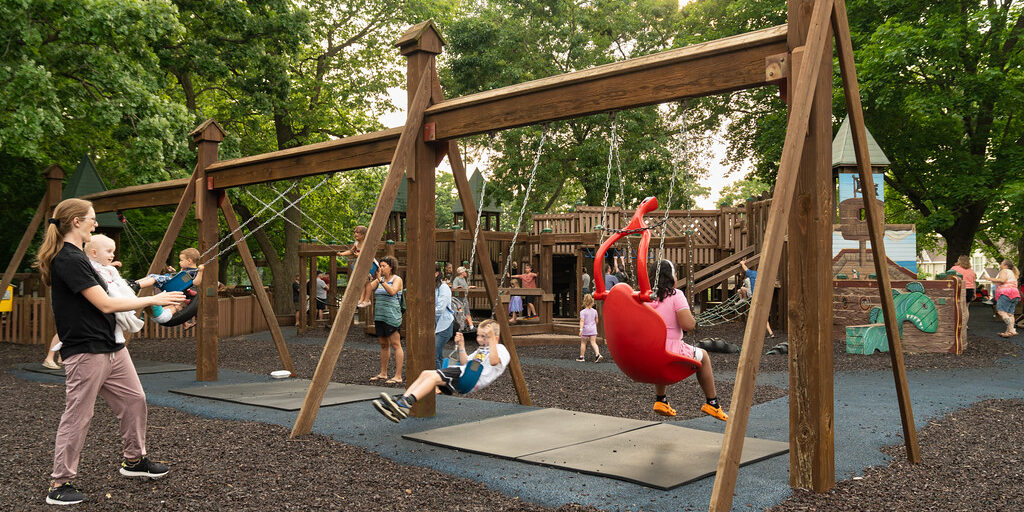 Children and adults are playing on swings and playground equipment in an outdoor park surrounded by trees on a sunny day.
