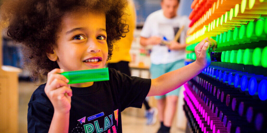 A child holds a translucent piece and interacts with a colorful light peg wall display in an indoor play area.