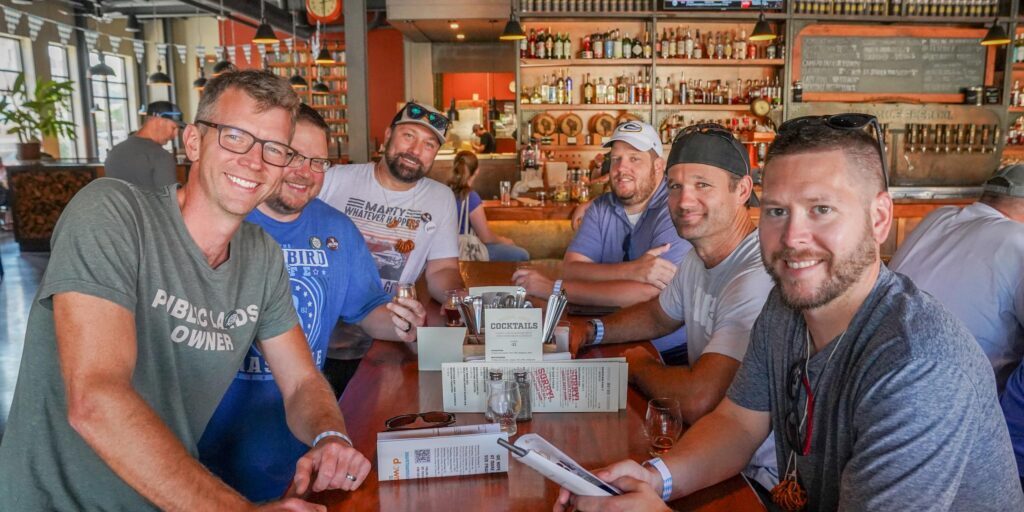 Six men sit around a wooden table at a bar, smiling at the camera. Menus and drinks are on the table; a TV with a sports game is visible in the background.