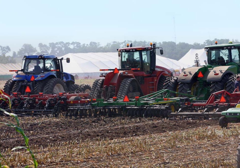 Three tractors—blue, red, and green—are lined up in a field with large farming equipment attached, with tents and trees visible in the background.