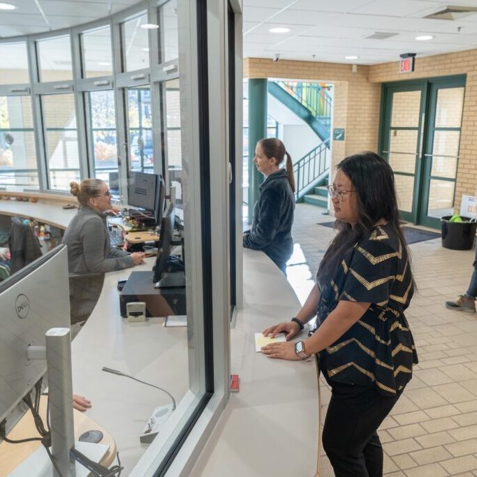 People stand in line at an indoor reception desk, speaking to staff members behind a glass partition in a brightly lit lobby.