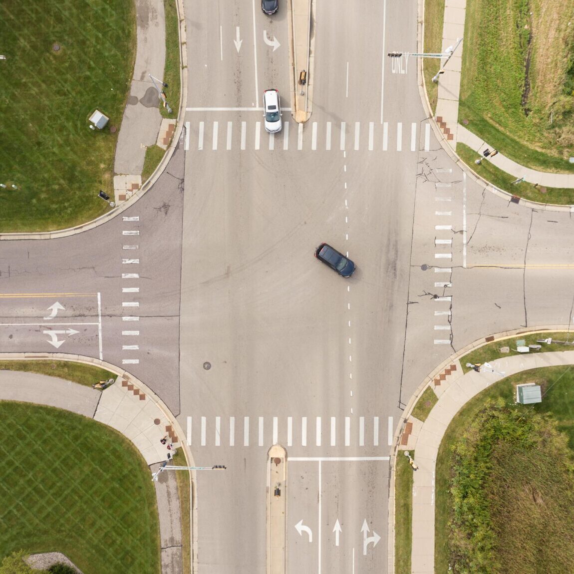 Aerial view of a four-way intersection with a single dark car in the center, surrounded by grass and multiple traffic lanes marked with arrows and crosswalks.