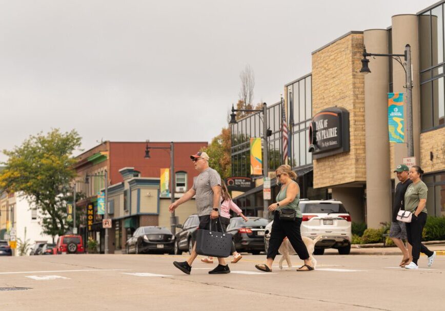 Four people walk across a downtown street crosswalk on a cloudy day, passing storefronts and parked cars.
