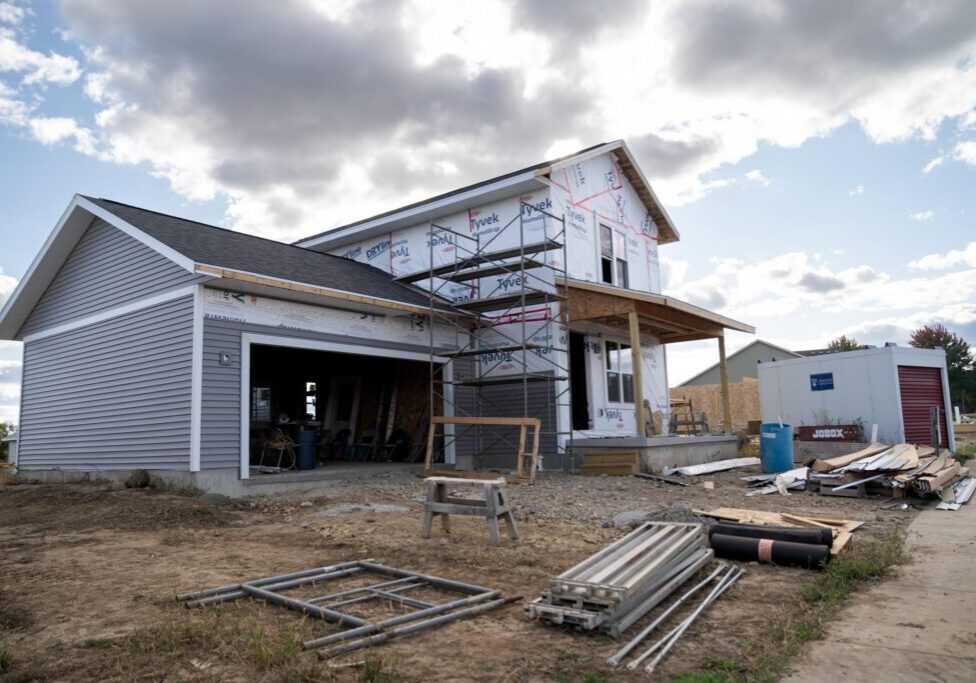 A two-story house under construction with scaffolding, exposed insulation, and building materials scattered around the unfinished yard.